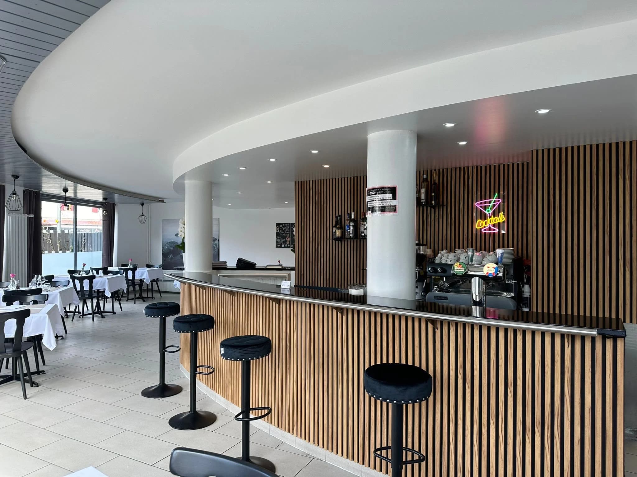 Le comptoir courbe en tasseaux de bois, néon Cocktails et machine à café, la salle dressée en arrière-plan