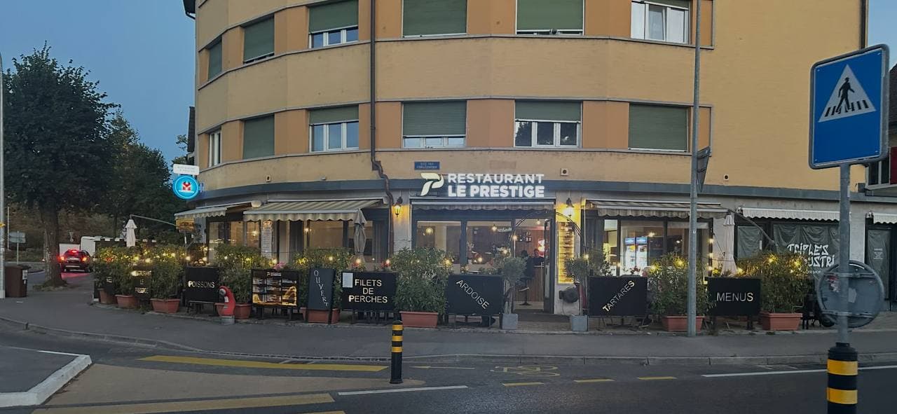 The front of Restaurant Le Prestige at dusk, the sign lit up on the street corner, the terrace and the menu boards